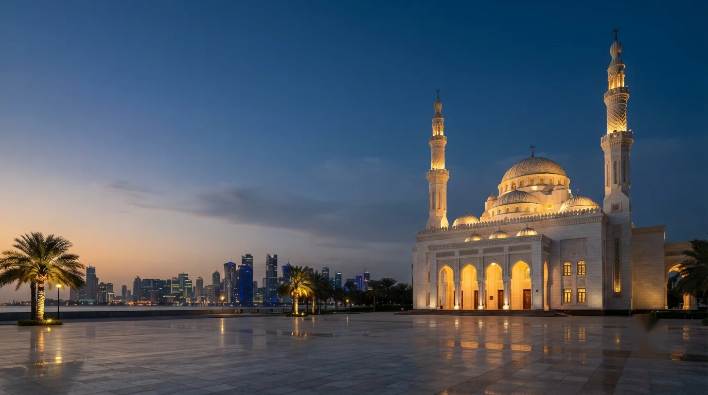 A white masjid with two minarets at dusk, a Gulf city skyline behind it